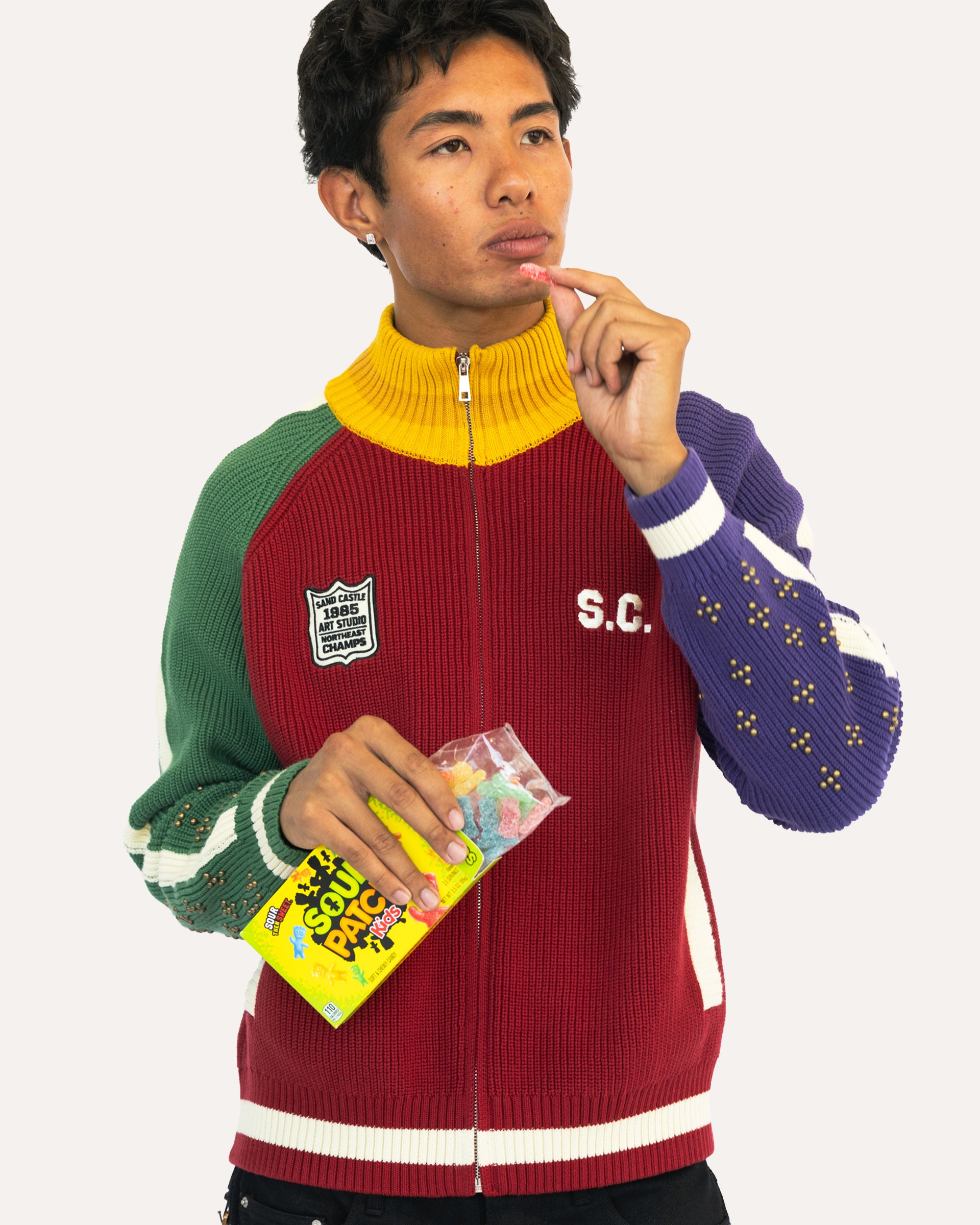 A man wearing a colorful ribbed zip-up sweater with green, yellow, red, and purple color-blocked sleeves and collar, holding a bag of Sour Patch Kids against a light gray background.